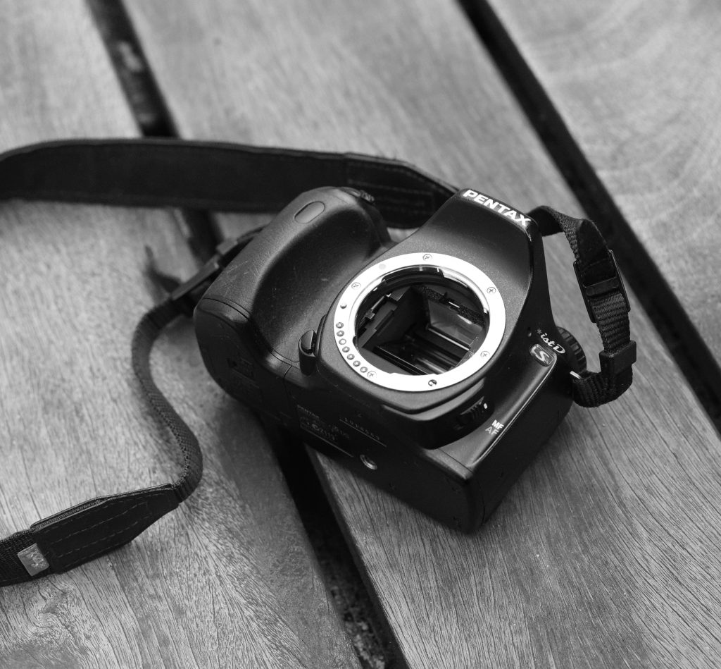 Black and white photograph of a black Pentax *ist DS DSLR camera body without a lens attached, resting on slatted wooden planks with its neck strap attached.