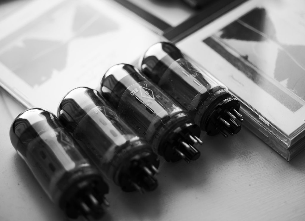 "Black-and-white photo: four Russian 6P3S-E vacuum tubes lying on their sides, pins facing the camera, beside CD editions of Bill Evans's 'The Paris Concert, Edition Two'."