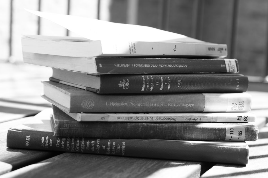 A stack of translated editions and the original Danish copy of Louis Hjelmslev's Omkring sprogteoriens grundlæggelse (Prolegomena to a Theory of Language, 1943), featuring book spines in Danish, English, French (Prolégomènes à une théorie du langage), German (Prolegomena zu einer Sprachtheorie), Italian (I fondamenti della teoria del linguaggio), Portuguese, and Spanish.
