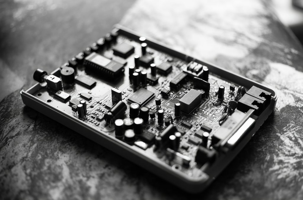 Black-and-white close-up photograph of an exposed circuit board resting on a mottled stone surface. The board fills most of the frame at an angle, its case opened to reveal densely packed components: numerous cylindrical electrolytic capacitors of varying heights, several rectangular integrated-circuit chips, a large square chip near the center, a boxy relay, and rows of soldered pins. Board silkscreen labels like "U26" and "C81" are visible. A shallow depth of field keeps the central components sharp while the near and far edges blur. This is the top side of a Trust 14k4 modem from the mid-1990s, similar to the hardware the NetLock BBS ran on.The SySop of NetLock BBS was Kasper Bergholt.